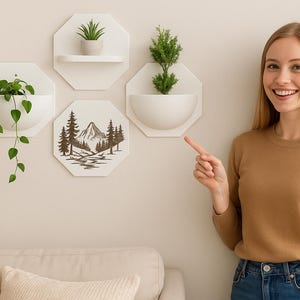 Peut inclure: Des jardinières murales octogonales blanches avec de la verdure et une scène de montagne. Une femme montre les jardinières, qui comprennent une petite étagère et une demi-cercle. Les plantes sont dans des pots blancs.