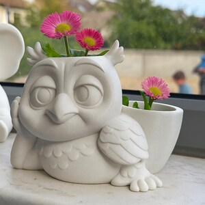 以下が含まれることがあります： ピンクの花と黄色の中心を持つ白いセラミック製のフクロウ型の植木鉢。フクロウは頭を手に乗せています。左側には別の白いセラミック象があります。植木鉢は窓枠にあります。