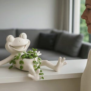 Könnte beinhalten: Ein skurriler, cremefarbener Keramik-Frosch-Pflanztopf mit ausgestreckten Armen und Beinen, der grüne Pflanzen hält. Der Frosch hat einen lächelnden Ausdruck. Der Pflanztopf steht auf einer weißen Oberfläche.