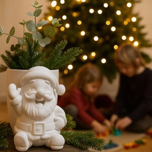 Könnte beinhalten: Eine weiße Keramikfigur des Weihnachtsmanns mit lächelndem Gesicht, Hut und Gürtel. Die Figur steht vor einer weißen Vase mit Grünzeug und einem Weihnachtsbaum mit Lichtern im Hintergrund. Festliche Weihnachtsdekoration.