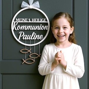 Könnte beinhalten: Ein weißer Holz-Kreis mit dem Text "Meine 1. Heilige Kommunion Pauline" und goldenen Fisch-Dekorationen, die daran hängen. Ein junges Mädchen in einem weißen Kleid steht vor dem Kreis.
