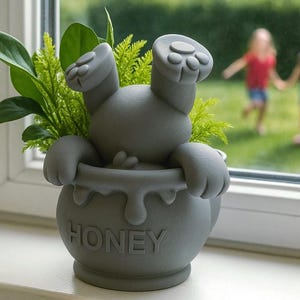 May include: A gray ceramic planter shaped like a bear, with its paws on the rim of a honey pot. The word "HONEY" is embossed on the pot. Green plants sprout from the pot. Two children play in the background.