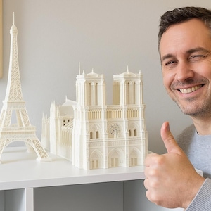 Könnte beinhalten: Elfenbeinfarbene 3D-gedruckte Modelle des Eiffelturms und der Kathedrale Notre-Dame. Das Eiffelturm-Modell befindet sich links, das Kathedralenmodell rechts. Ein Mann lächelt und gibt ein Daumen hoch.