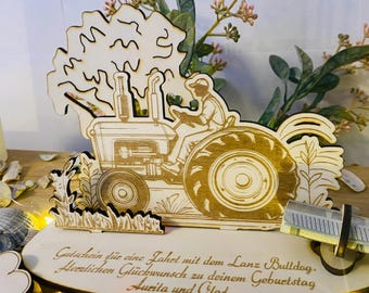 May include: A wooden tractor with a figure of a person driving it. The tractor is surrounded by green grass and a tree. The tractor is a gift certificate holder with the text "Gutschein fue eine Fahrt mit dem Lanz Bulldog. Herzlichen Glückwunsch zu deinem Geburtstag Aurita und Clad" on the base.