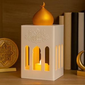 Puede incluir: Una linterna blanca con cortes arqueados y una cúpula dorada, iluminada por una vela en el interior. Una luna creciente dorada y un disco decorativo dorado en forma de estrella están a cada lado. Libros en el fondo.