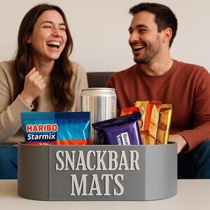 May include: A gray snack container with the text "Snackbar Mats" on the front. The container is filled with various snacks, including a blue bag of Haribo Starmix candy, a silver can, a purple chocolate bar, and a gold-wrapped chocolate bar.