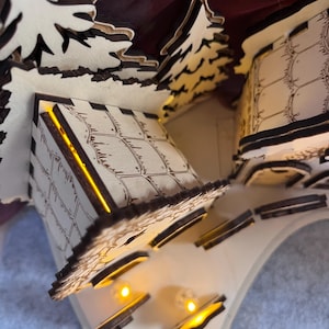 K&ouml;nnte beinhalten: Eine lasergeschnittene Holzdorf-Szene mit zwei kleinen Geb&auml;uden und immergr&uuml;nen B&auml;umen. Die Strukturen haben ge&auml;tzte Gittermuster an den W&auml;nden und werden von innen durch warmgelbe LED-Lichter beleuchtet, die einen sanften Schein abgeben.