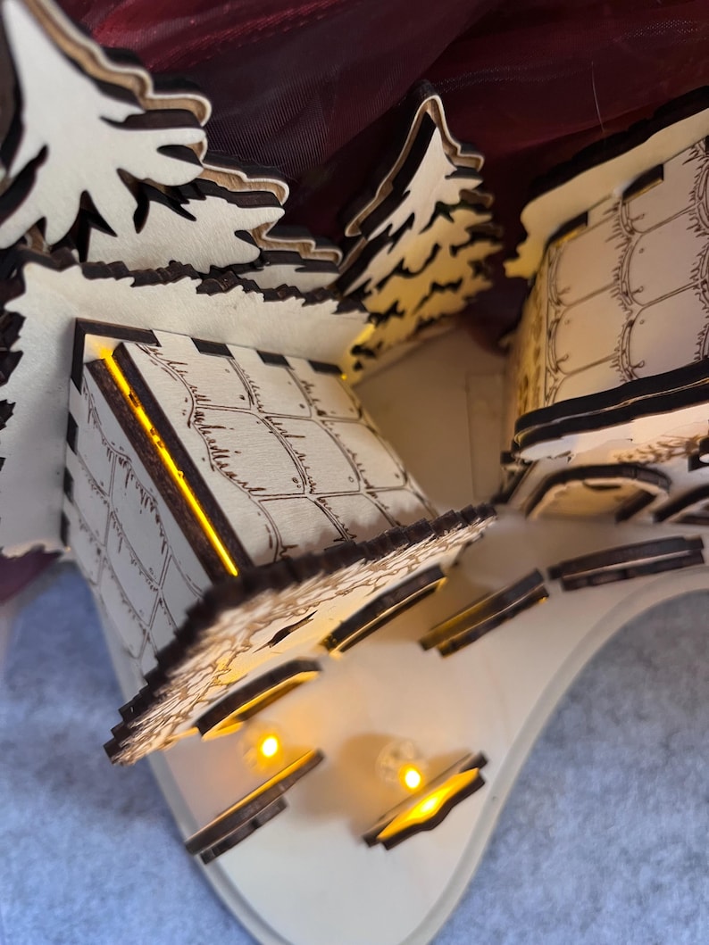 K&ouml;nnte beinhalten: Eine lasergeschnittene Holzdorf-Szene mit zwei kleinen Geb&auml;uden und immergr&uuml;nen B&auml;umen. Die Strukturen haben ge&auml;tzte Gittermuster an den W&auml;nden und werden von innen durch warmgelbe LED-Lichter beleuchtet, die einen sanften Schein abgeben.