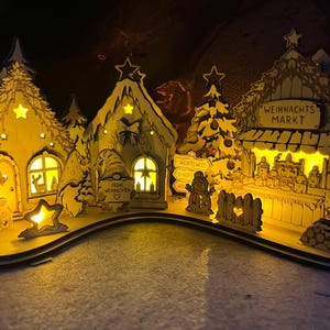 K&ouml;nnte beinhalten: Ein lasergeschnittenes Weihnachtsdorf aus Holz mit beleuchteten H&auml;usern, Tannenb&auml;umen und Wichteln. Die Szene zeigt einen Stand mit der Aufschrift WEIHNACHTS MARKT, ein Schild mit WICHTELWELT und einen Wichtel mit dem Schild FROHE WEIHNACHTEN.