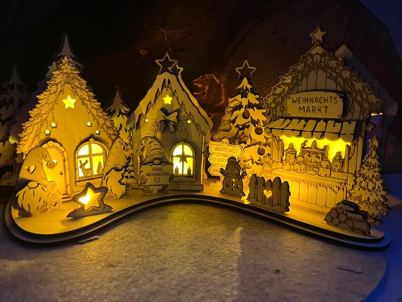 K&ouml;nnte beinhalten: Ein lasergeschnittenes Weihnachtsdorf aus Holz mit beleuchteten H&auml;usern, Tannenb&auml;umen und Wichteln. Die Szene zeigt einen Stand mit der Aufschrift WEIHNACHTS MARKT, ein Schild mit WICHTELWELT und einen Wichtel mit dem Schild FROHE WEIHNACHTEN.
