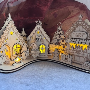 K&ouml;nnte beinhalten: Ein weihnachtliches Dorf aus Holz mit beleuchteten H&auml;usern, Tannenb&auml;umen und Wichteln. Die Szene enth&auml;lt einen Stand mit der Aufschrift "WEIHNACHTS MARKT" und ein Schild mit "FROHE WEIHNACHTEN". Die Strukturen bestehen aus hellem Holz mit Ausschnitten.