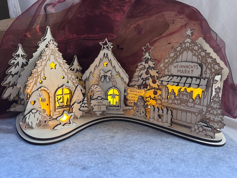 K&ouml;nnte beinhalten: Ein weihnachtliches Dorf aus Holz mit beleuchteten H&auml;usern, Tannenb&auml;umen und Wichteln. Die Szene enth&auml;lt einen Stand mit der Aufschrift "WEIHNACHTS MARKT" und ein Schild mit "FROHE WEIHNACHTEN". Die Strukturen bestehen aus hellem Holz mit Ausschnitten.