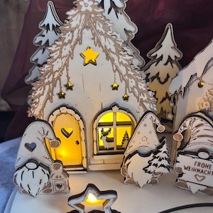 K&ouml;nnte beinhalten: Ein beleuchtetes Weihnachtsdorf aus Holz mit einem Haus, das gelb leuchtet, einer herzf&ouml;rmigen T&uuml;r und Sternausschnitten. Drei Wichtelfiguren stehen davor, eine davon h&auml;lt ein Schild mit der Aufschrift "FROHE WEIHNACHTEN" und einem Herz.