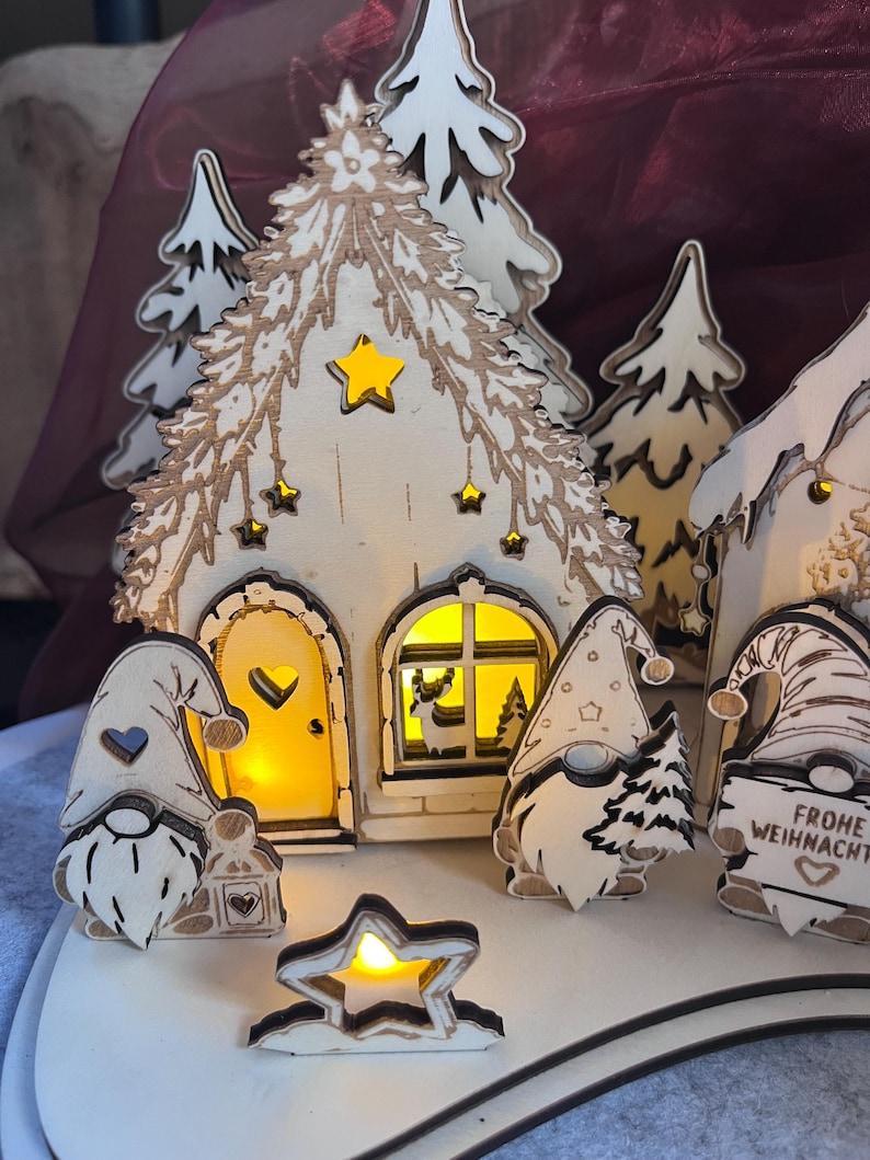 K&ouml;nnte beinhalten: Ein beleuchtetes Weihnachtsdorf aus Holz mit einem Haus, das gelb leuchtet, einer herzf&ouml;rmigen T&uuml;r und Sternausschnitten. Drei Wichtelfiguren stehen davor, eine davon h&auml;lt ein Schild mit der Aufschrift "FROHE WEIHNACHTEN" und einem Herz.