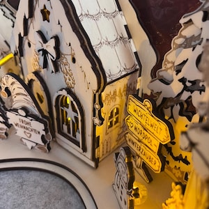 K&ouml;nnte beinhalten: Eine lasergeschnittene Weihnachtsdorf-Szene aus Holz mit einem kleinen Haus mit beleuchteten Fenstern, einem Wichtel, der ein Schild mit der Aufschrift FROHE WEIHNACHTEN h&auml;lt, und einem Wegweiser mit dem Text WICHTELWELT, NORDPOL und WINTERZAUBER.