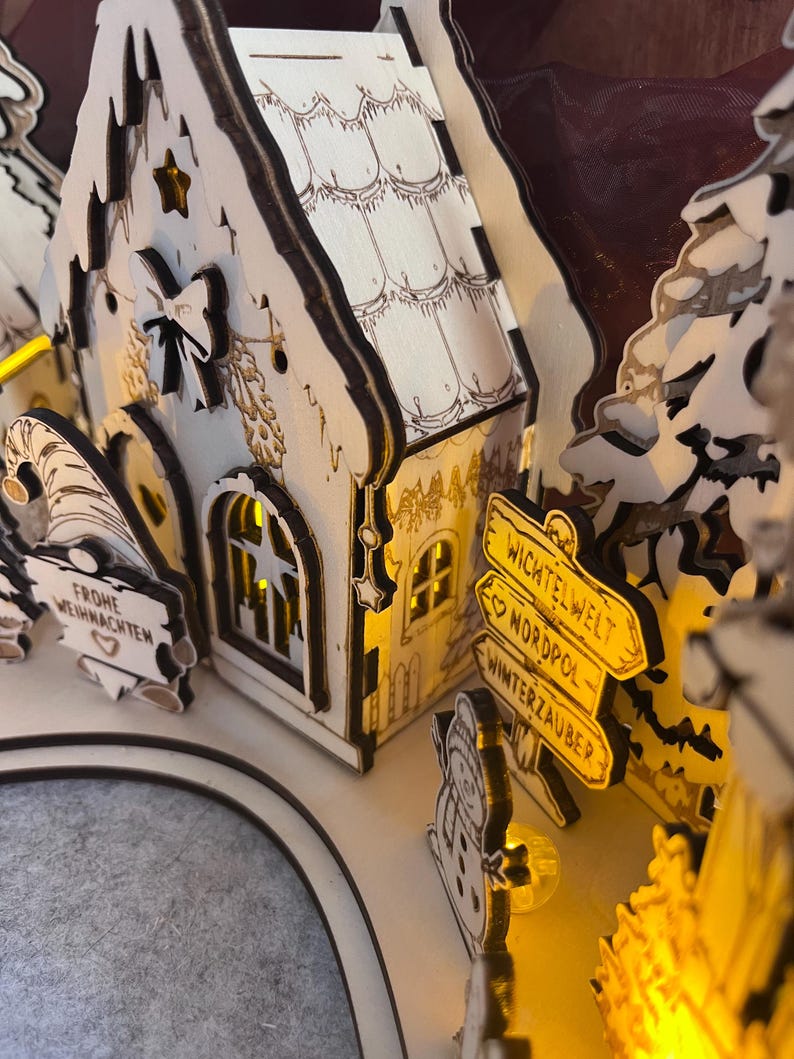K&ouml;nnte beinhalten: Eine lasergeschnittene Weihnachtsdorf-Szene aus Holz mit einem kleinen Haus mit beleuchteten Fenstern, einem Wichtel, der ein Schild mit der Aufschrift FROHE WEIHNACHTEN h&auml;lt, und einem Wegweiser mit dem Text WICHTELWELT, NORDPOL und WINTERZAUBER.