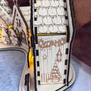 K&ouml;nnte beinhalten: Ein lasergeschnittener Weihnachtsdorf-Bogen aus hellem Holz mit dunkel gebrannten Kanten. Die Struktur umfasst ein Haus mit Schindeldach und Seitenw&auml;nden, die mit einer Laterne, einem Weihnachtsbaum und festlichen Girlanden graviert sind.