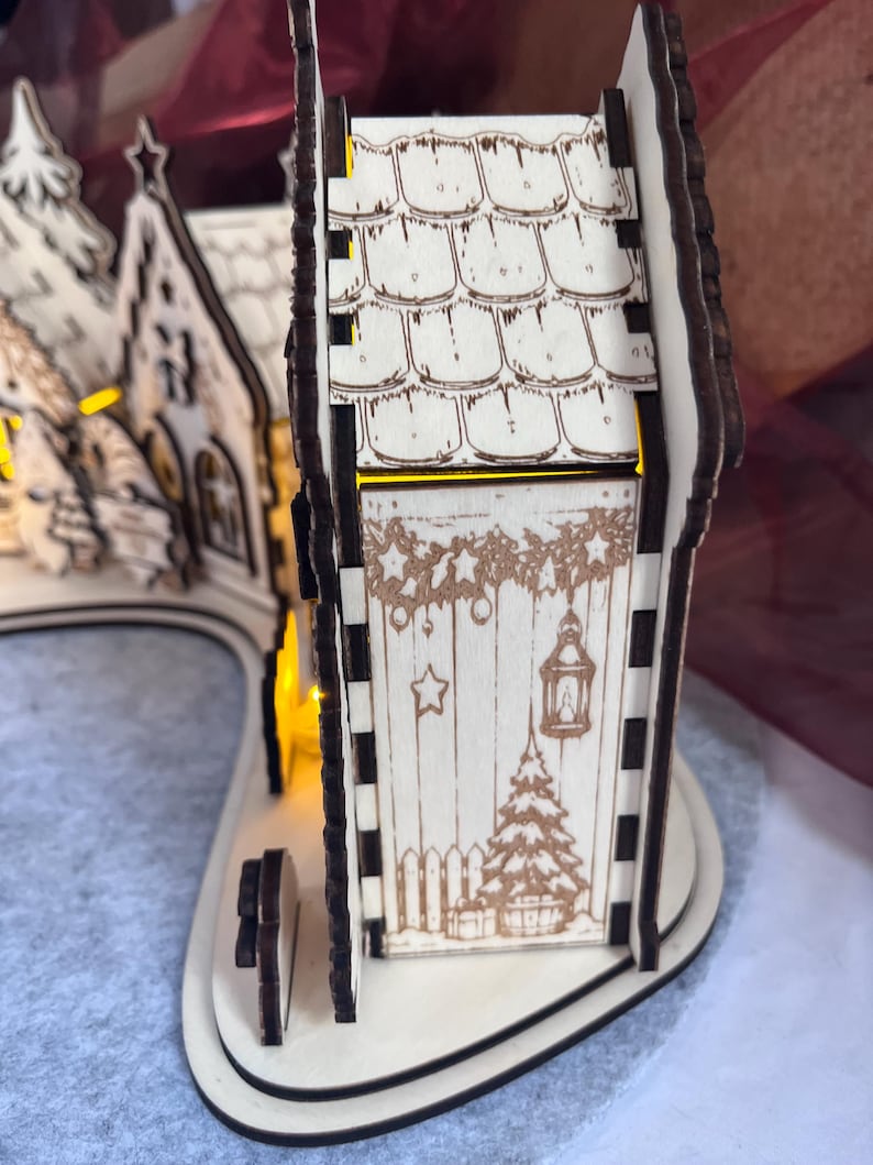 K&ouml;nnte beinhalten: Ein lasergeschnittener Weihnachtsdorf-Bogen aus hellem Holz mit dunkel gebrannten Kanten. Die Struktur umfasst ein Haus mit Schindeldach und Seitenw&auml;nden, die mit einer Laterne, einem Weihnachtsbaum und festlichen Girlanden graviert sind.