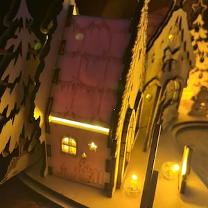 K&ouml;nnte beinhalten: Ein lasergeschnittenes Miniaturhaus aus Holz mit rosafarbenem Dach und W&auml;nden, das von innen durch warmgelbe LED-Lichter beleuchtet wird. Die Struktur zeigt ausgeschnittene Fenster, Sternformen und Baum-Silhouetten in einer leuchtenden Schichtoptik.