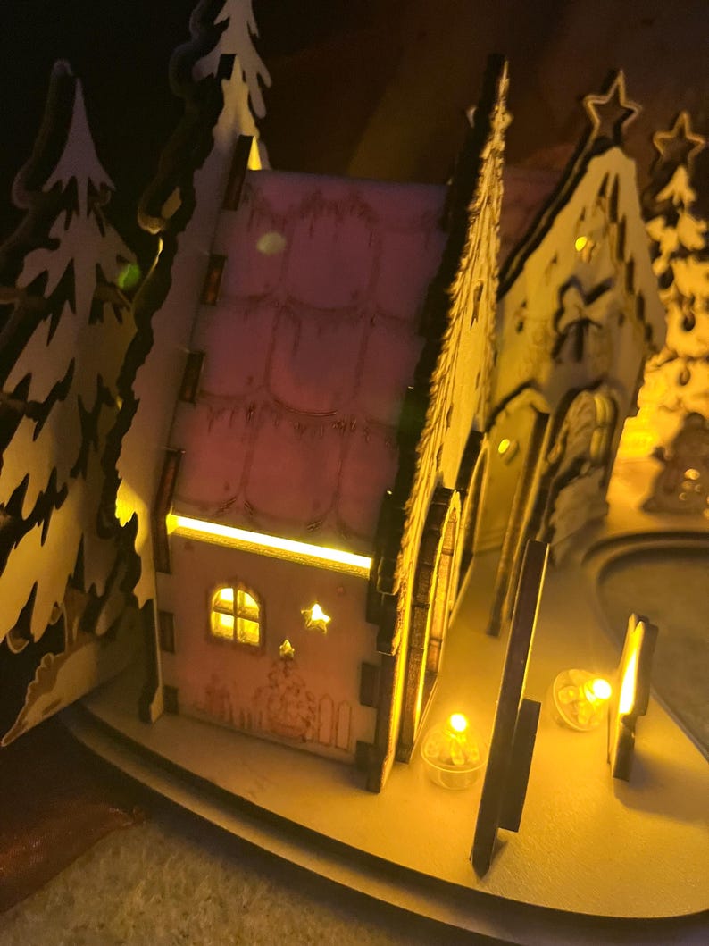 K&ouml;nnte beinhalten: Ein lasergeschnittenes Miniaturhaus aus Holz mit rosafarbenem Dach und W&auml;nden, das von innen durch warmgelbe LED-Lichter beleuchtet wird. Die Struktur zeigt ausgeschnittene Fenster, Sternformen und Baum-Silhouetten in einer leuchtenden Schichtoptik.