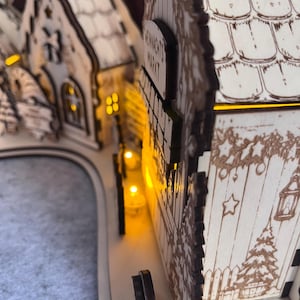 K&ouml;nnte beinhalten: Ein dekorativer Holzbogen in heller Farbe mit einer Miniatur-Dorflandschaft und lasergeschnittenen Details. Die Struktur umfasst kleine Geb&auml;ude mit Ziegeld&auml;chern, Sternmotiven und einem Schild mit der Aufschrift WEIHNACHTS MARKT. Warme gelbe LED-Lichter beleuchten das Innere.