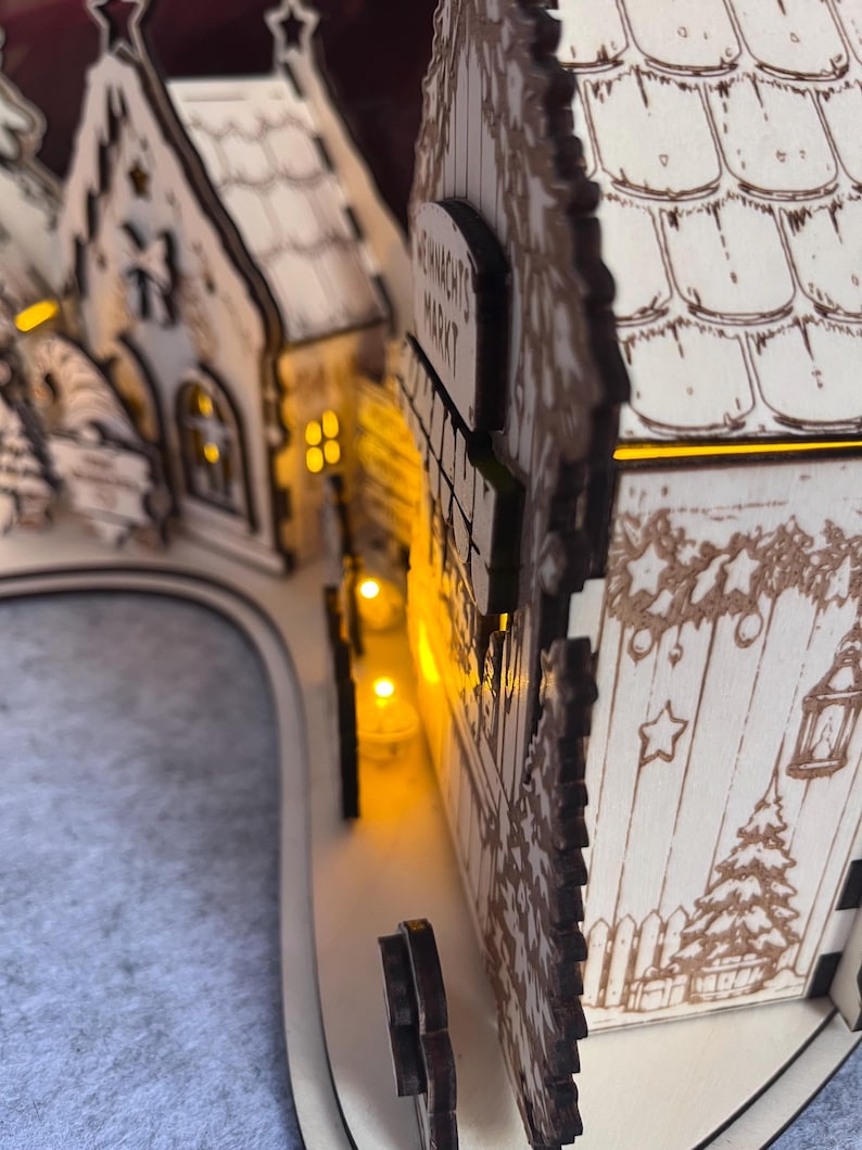 K&ouml;nnte beinhalten: Ein dekorativer Holzbogen in heller Farbe mit einer Miniatur-Dorflandschaft und lasergeschnittenen Details. Die Struktur umfasst kleine Geb&auml;ude mit Ziegeld&auml;chern, Sternmotiven und einem Schild mit der Aufschrift WEIHNACHTS MARKT. Warme gelbe LED-Lichter beleuchten das Innere.