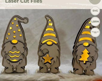 May include: Three laser-cut wooden gnome figures with internal warm yellow lighting. The left gnome holds a round ornament, the center gnome holds a star, and the right gnome holds a Christmas tree. Text includes EY ART, Laser Cut Files, 3-4 mm optimized, SVG, LBRN, and Private use only.