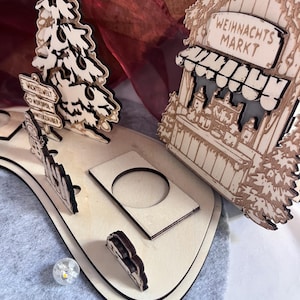K&ouml;nnte beinhalten: Ein lasergeschnittenes Weihnachts-Dorf-Display aus Holz mit einem Baum, einem Wegweiser mit dem Text WICHTELWELT, NORDPOL und WINTERZAUBER sowie einem Marktstand mit dem Text WEIHNACHTS MARKT. Die Basis enth&auml;lt kreisf&ouml;rmige Aussparungen f&uuml;r Lichter.