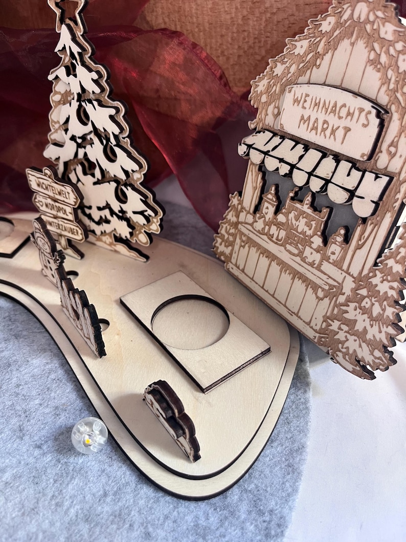 K&ouml;nnte beinhalten: Ein lasergeschnittenes Weihnachts-Dorf-Display aus Holz mit einem Baum, einem Wegweiser mit dem Text WICHTELWELT, NORDPOL und WINTERZAUBER sowie einem Marktstand mit dem Text WEIHNACHTS MARKT. Die Basis enth&auml;lt kreisf&ouml;rmige Aussparungen f&uuml;r Lichter.