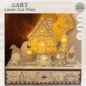 Könnte beinhalten: Ein lasergeschnittenes Holzdisplay mit einem Haus, zwei Gnomen, einem Briefkasten und einer Laterne, beleuchtet durch warmgelbes Licht. Der Sockel zeigt eingravierte Illustrationen einer Truhe, einer Laterne und einer Schubkarre mit dem Text Wichielschatz.