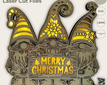 Könnte beinhalten: Eine lasergeschnittene Holzdekoration mit drei Gnomen mit hohen Mützen, die jeweils einzigartige Muster und Ausschnitte aufweisen. Der mittlere Gnom hält ein Schild mit der Aufschrift MERRY CHRISTMAS und zwei kleinen Baum-Symbolen.