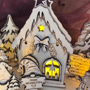 K&ouml;nnte beinhalten: Eine wei&szlig;e Holzhaus-Dekoration mit Sternausschnitt an der Spitze, gelbem Sternakzent und Schleife. Ein Wichtel davor h&auml;lt ein Schild mit der Aufschrift FROHE WEIHNACHTEN. Ein gelb beleuchtetes Fenster ist sichtbar, neben einem Wegweiser WICHTELWELT.
