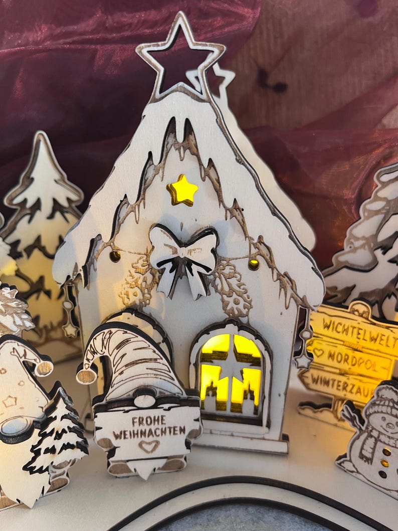 K&ouml;nnte beinhalten: Eine wei&szlig;e Holzhaus-Dekoration mit Sternausschnitt an der Spitze, gelbem Sternakzent und Schleife. Ein Wichtel davor h&auml;lt ein Schild mit der Aufschrift FROHE WEIHNACHTEN. Ein gelb beleuchtetes Fenster ist sichtbar, neben einem Wegweiser WICHTELWELT.