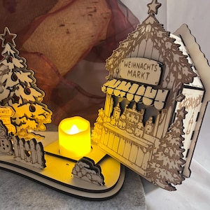 K&ouml;nnte beinhalten: Eine lasergeschnittene Weihnachtsmarktszene aus Holz mit einem kleinen Stand mit der Aufschrift WEIHNACHTS MARKT, einem geschm&uuml;ckten Baum, einem Schneemann und einem Zaun. Ein batteriebetriebenes gelbes LED-Teelicht steht in der Mitte.