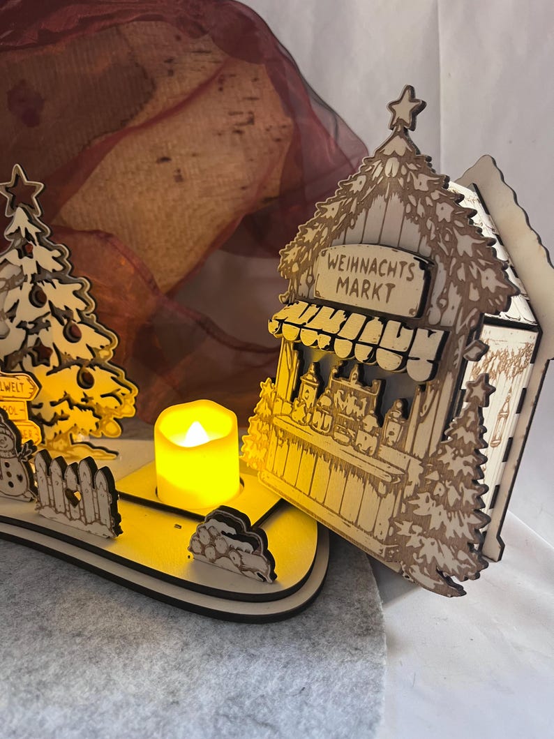 K&ouml;nnte beinhalten: Eine lasergeschnittene Weihnachtsmarktszene aus Holz mit einem kleinen Stand mit der Aufschrift WEIHNACHTS MARKT, einem geschm&uuml;ckten Baum, einem Schneemann und einem Zaun. Ein batteriebetriebenes gelbes LED-Teelicht steht in der Mitte.