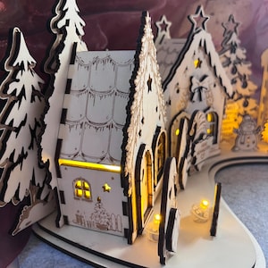 K&ouml;nnte beinhalten: Ein lasergeschnittenes Weihnachtsdorf aus Holz mit zwei H&auml;usern mit Bogent&uuml;ren, schneebedeckten D&auml;chern und Tannenb&auml;umen. Die Struktur wird von innen durch warmgelbe LED-Lichter beleuchtet, die einen Schein durch die Fenster und sternf&ouml;rmigen Ausschnitte werfen.
