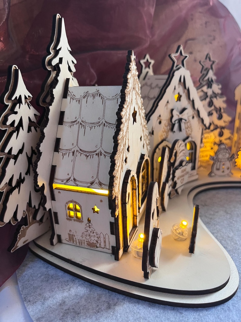 K&ouml;nnte beinhalten: Ein lasergeschnittenes Weihnachtsdorf aus Holz mit zwei H&auml;usern mit Bogent&uuml;ren, schneebedeckten D&auml;chern und Tannenb&auml;umen. Die Struktur wird von innen durch warmgelbe LED-Lichter beleuchtet, die einen Schein durch die Fenster und sternf&ouml;rmigen Ausschnitte werfen.