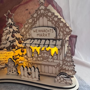 K&ouml;nnte beinhalten: Eine lasergeschnittene Weihnachtsmarktszene aus Holz mit einem Stand mit der Aufschrift WEIHNACHTS MARKT, einem Schneemann, einem Wegweiser mit der Beschriftung WICHTELWELT, NORDPOL und WINTER sowie Tannenb&auml;umen. Die Szene wird von innen beleuchtet.