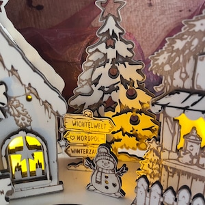 K&ouml;nnte beinhalten: Ein dekoratives Weihnachtsdorf-Set aus Holz mit beleuchteten wei&szlig;en H&auml;usern, einem stilisierten Baum und einem Schneemann. Ein Wegweiser zeigt die Aufschrift WICHTELWELT, NORDPOL und WINTERZAUBER. Die Teile haben dunkle Kanten und warmes Licht.