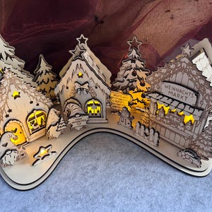 K&ouml;nnte beinhalten: Ein helles Holz-Weihnachtsdorf mit beleuchteten H&auml;usern, Tannenb&auml;umen und Wichteln. Die Geb&auml;ude tragen Schilder mit der Aufschrift "Weihnachts Markt", "Wichtelwelt" und "Frohe Weihnachten". Die Szene ist auf einer geschwungenen Basis angeordnet.