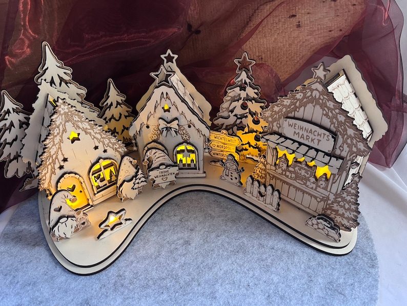 K&ouml;nnte beinhalten: Ein helles Holz-Weihnachtsdorf mit beleuchteten H&auml;usern, Tannenb&auml;umen und Wichteln. Die Geb&auml;ude tragen Schilder mit der Aufschrift "Weihnachts Markt", "Wichtelwelt" und "Frohe Weihnachten". Die Szene ist auf einer geschwungenen Basis angeordnet.