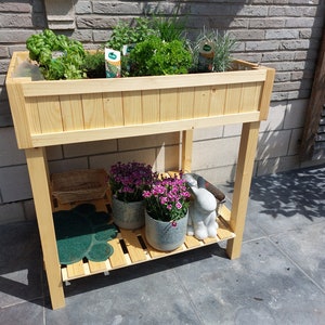 Op de afbeelding: Een houten plantenstandaard met twee planken. De bovenste plank heeft een plantenbak gevuld met groene kruiden. De onderste plank heeft twee planten in potten met roze bloemen, een klein decoratief konijn en een groene mat.
