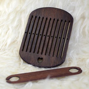 May include: A dark brown wooden weaving comb with brass rods and a heart-shaped cutout. A smaller, matching shuttle lies below. The comb is approximately 5 inches wide, and the shuttle is about 7 inches long. The items are displayed on a white, fluffy surface.