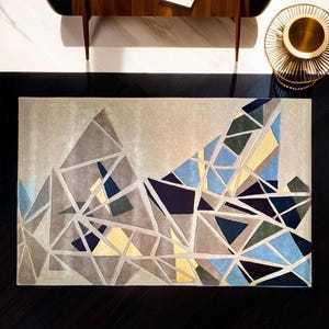 May include: Rectangular rug with a geometric design. The rug has a light beige background with a pattern of interconnected triangles in shades of grey, blue, yellow, and green. The rug is placed on a dark wooden floor.