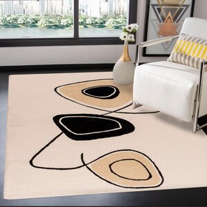 Puede incluir: Alfombra beige con un diseño moderno. La alfombra presenta tres formas abstractas grandes en negro y beige, delineadas con líneas negras. La alfombra está colocada en un salón con una silla blanca y un jarrón con flores.