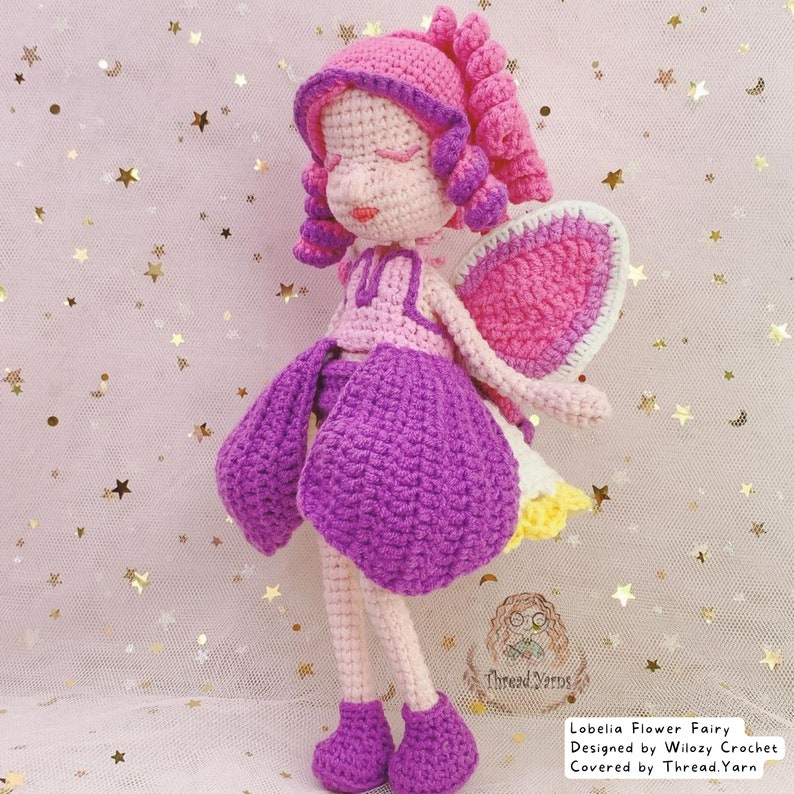 May include: A crocheted fairy doll with pink hair, purple dress, and pink wings. The doll is standing on a pink background with gold stars.