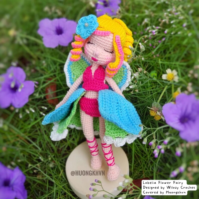 May include: A crocheted fairy doll with a pink dress, blue wings, and yellow hair. The doll is sitting on a wooden base and surrounded by purple flowers.