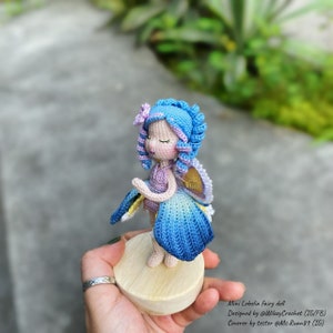 May include: A crocheted fairy doll with blue hair, a purple dress, and blue wings. The doll is standing on a wooden base.