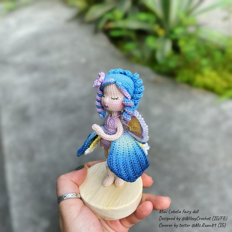 May include: A crocheted fairy doll with blue hair, a purple dress, and blue wings. The doll is standing on a wooden base.