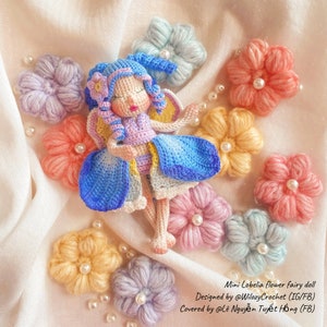 May include: A crocheted fairy doll with blue and purple clothing, sleeping on a bed of colorful crocheted flowers. The doll has a blue and purple dress, blue wings, and a purple flower in her hair. The flowers are in shades of pink, yellow, and blue.